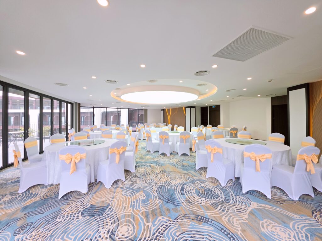 Phòng tiệc Ballroom 2 tại Soc Village setup bàn tròn sang trọng với nơ ghế vàng nhạt và thảm hoa văn xanh.